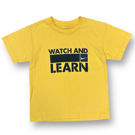 A yellow short sleeve Nike t-shirt with the text 'Watch and Learn' printed in black.