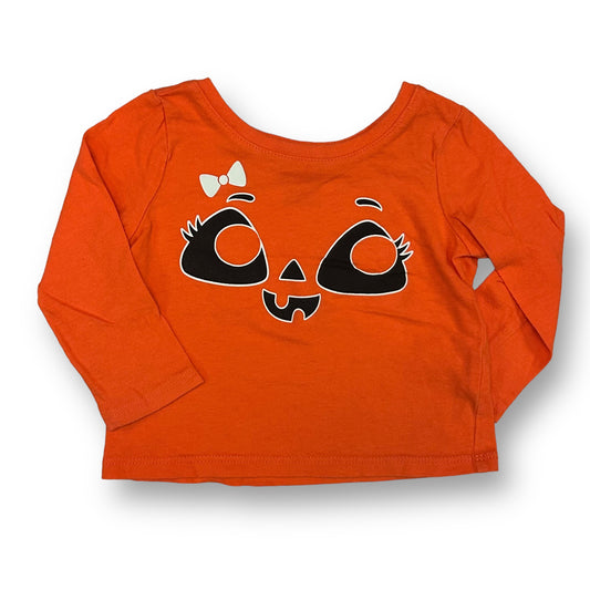 An orange children's shirt with a character face design resembling a pumpkin, ghost, or skeleton, and a bow detail.